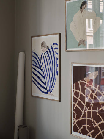 The Poster Clubs Sofia Lind - Blue Stripe At Concorde (50x70) Giclée print hangs on a light gray wall, showing a partially obscured figure among bold blue, beige, and brown shapes; a rolled paper tube rests below.