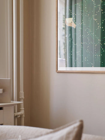 A softly lit, minimal corner with beige walls features part of The Poster Clubs Sofia Lind - Coffee Alone At Place De Clichy (30x40) giclée print, showing a green abstract design beside a white cabinet and exposed pipes.