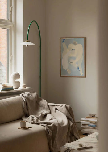 A cozy, minimalist living room with a beige sofa, a throw blanket, a cup and saucer, stacked books, soft natural light, and The Poster Clubs Sofia Lind - Hold You - Blue (50x70) art print displayed on the wall.