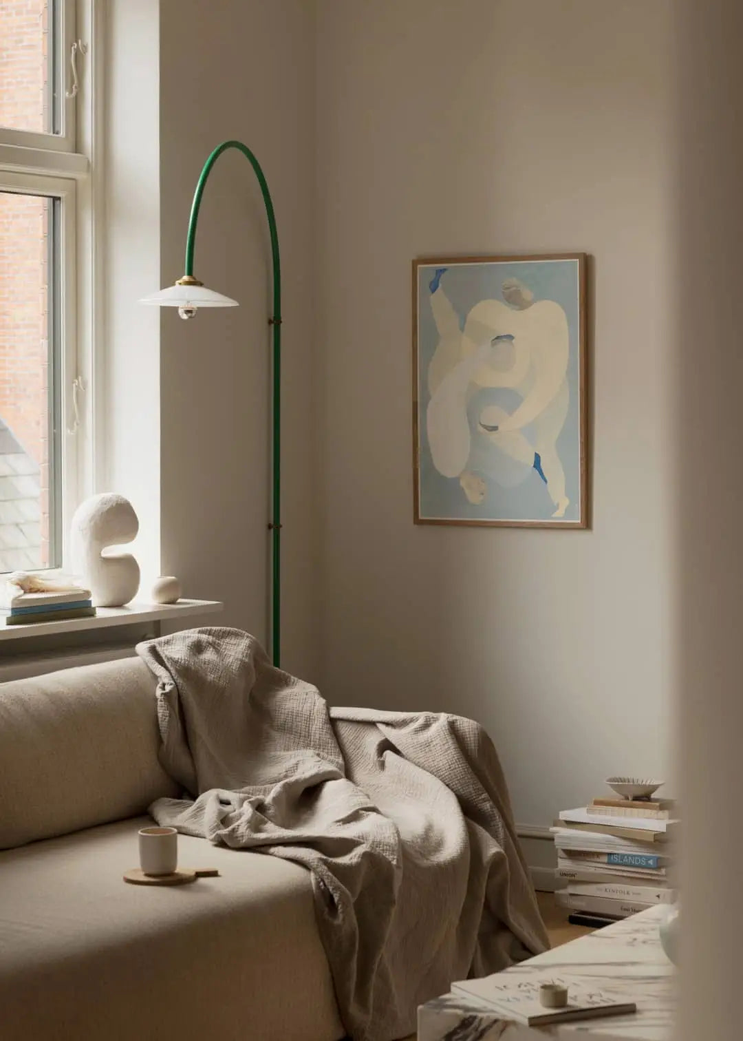 A cozy, minimalist living room with a beige sofa, blanket, books, sculptural decor, and a green floor lamp. On the wall is The Poster Club Berit Mogensen Lopez - Checkered (50x70) art print, illuminated by warm natural light.