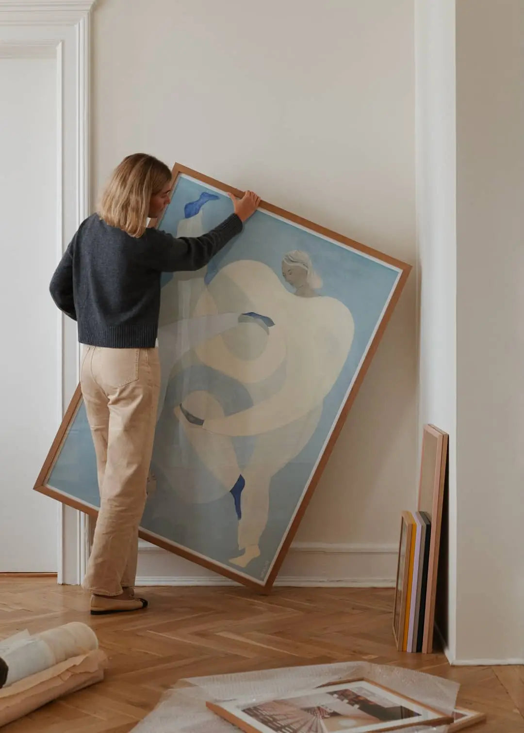 A person with shoulder-length hair, in a dark sweater and beige pants, places The Poster Club Berit Mogensen Lopez - Improvise (50x70) print with blue and beige tones against a wall in a minimalist room with wooden flooring and rolled-up art nearby.
