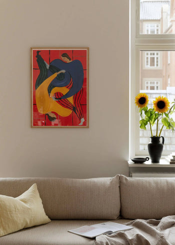 A cozy living room features a beige sofa, yellow pillow, and magazine. On the wall hangs Sofia Lind - Hold You - Red (30x40) from The Poster Club; two sunflowers in a black vase sit on the windowsill beside a cup, brightened by daylight.