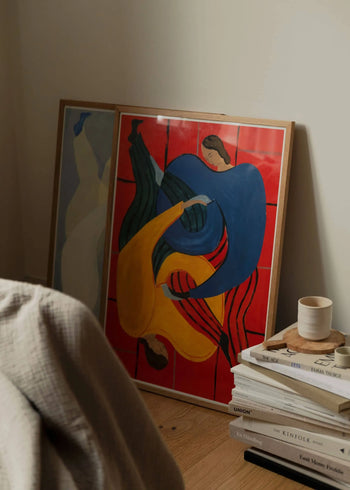 The Poster Clubs Sofia Lind - Hold You - Red (30x40) framed art print leans against the wall, its abstract blue and yellow forms standing out. Nearby, a stack of books and a ceramic cup rest on a wooden table.