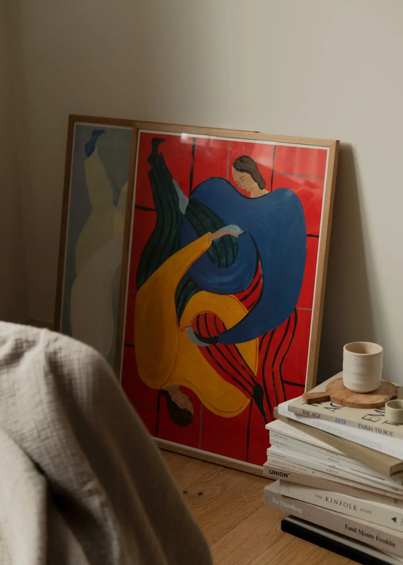 The Poster Club’s Sofia Lind - Hold You - Red (50x70) framed print leans stylishly against a wall, beside books, a cup, and a beige blanket—a scene echoing The Poster Club’s signature curated aesthetic.