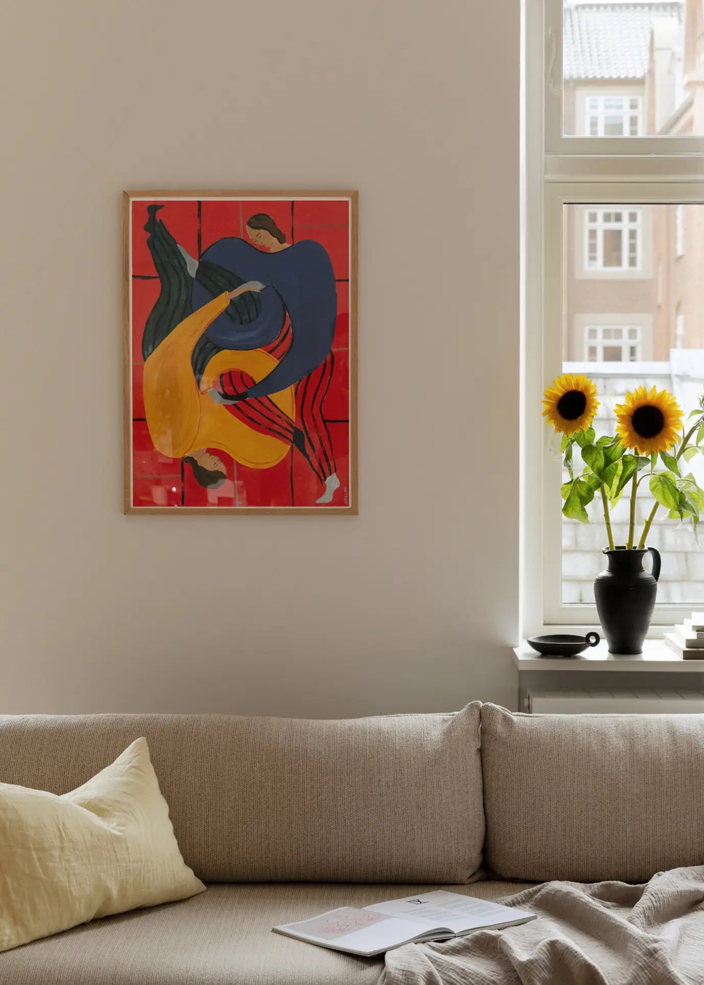 A modern living room with a beige sofa, yellow pillow, magazine, and The Poster Club Sofia Lind - Hold You - Red (50x70) art print on the wall. On the window ledge are a vase with two sunflowers and a cup.