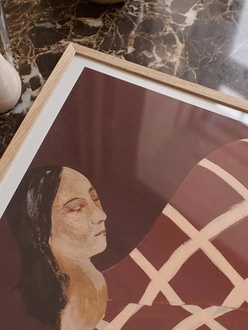 The Poster Clubs Berit Mogensen Lopez - Checkered (50x70) framed art print, featuring a stylized woman with long dark hair and closed eyes on a brown background with beige lines, is displayed on a marble surface.