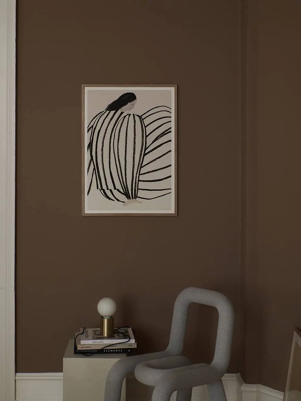 A minimal modern room with brown walls features the Improvise (50x70) art print by Berit Mogensen Lopez from The Poster Club, a sculptural light gray chair, a book-topped side table, and a round bulb lamp with a gold base.