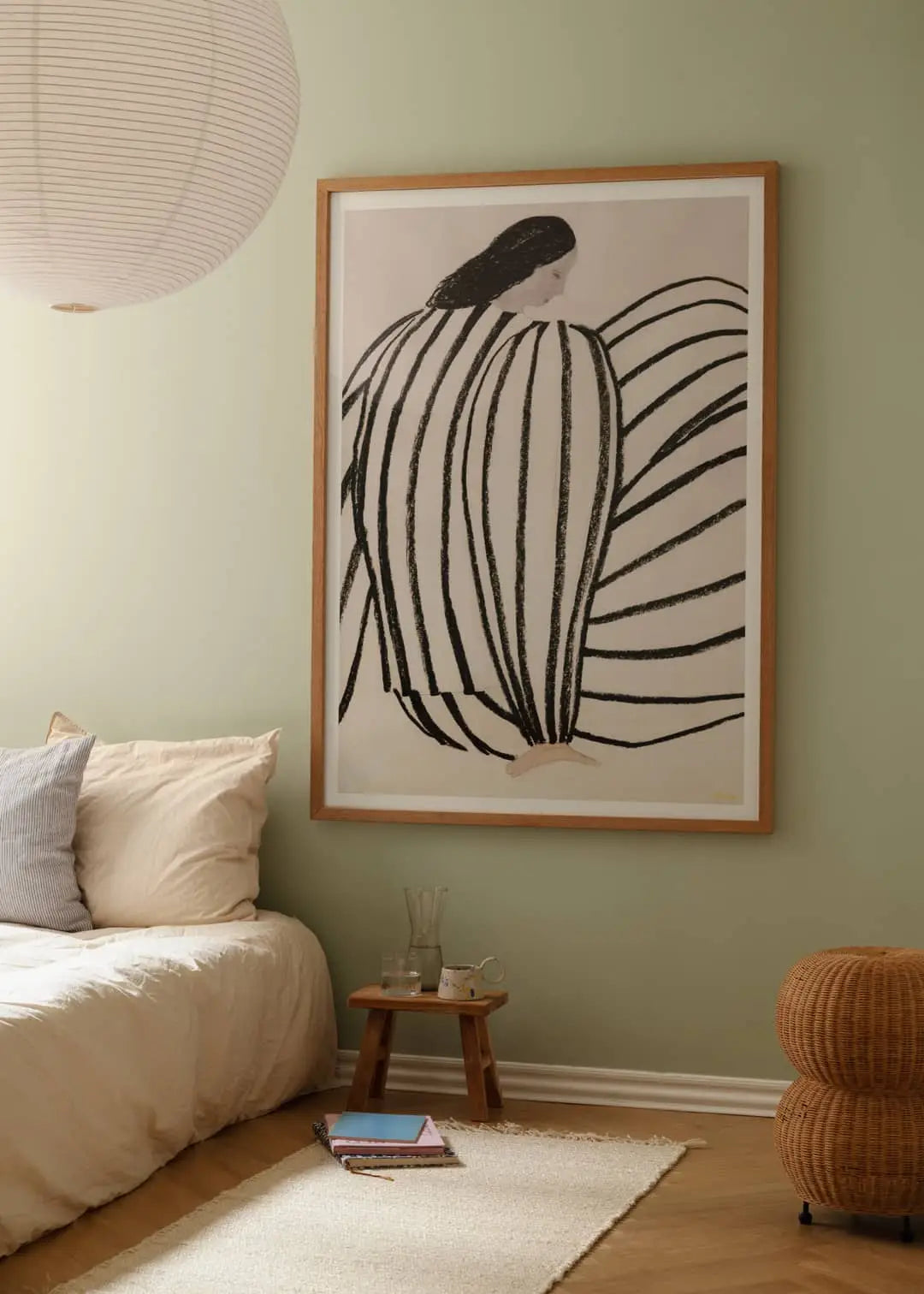A minimal, cozy bedroom features a low bed with beige bedding, a small wooden side table, and The Poster Club Berit Mogensen Lopez - Checkered (50x70) art print. Overhead, a round paper lantern gives the calming space a soft glow.