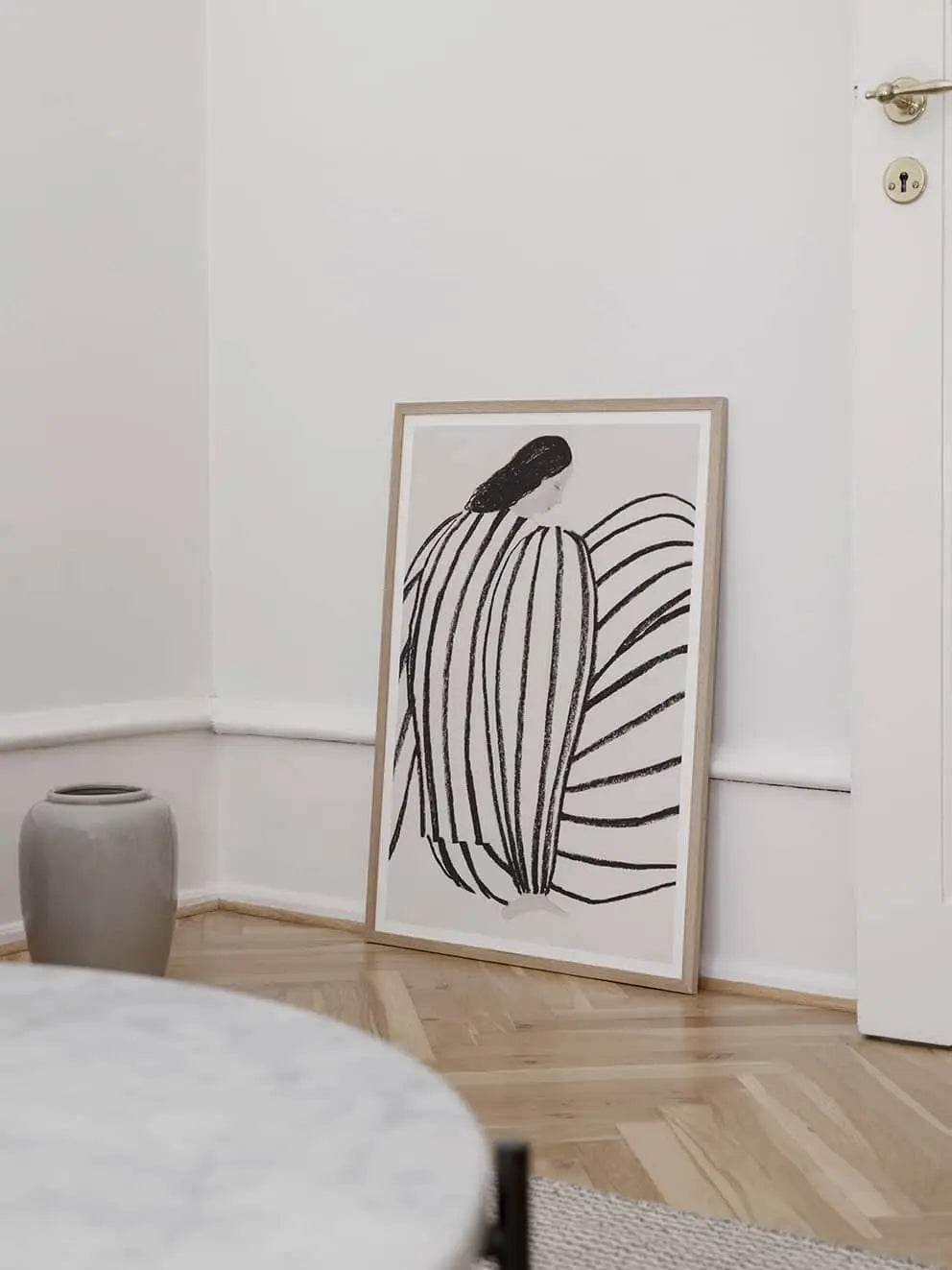 The Poster Club Berit Mogensen Lopez - Checkered (50x70), a framed abstract black-and-white art print, leans against the wall in a minimalist room beside a gray vase and round marble table—an elegant piece from The Poster Club.