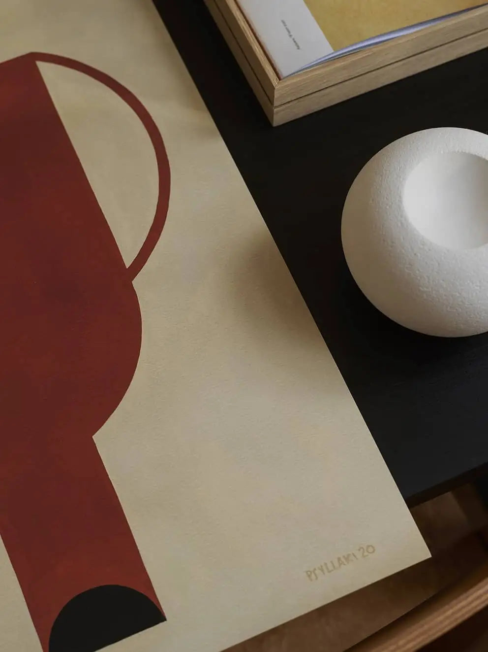 The Poster Club Studio Paradissi - Dialogues 03 (50x70): Minimalist abstract art print with red and black geometric shapes on beige, placed on a dark table with a round white textured object and a closed book. Printed on premium art paper. Brand: The Poster Club.