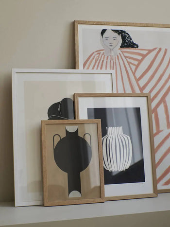Four framed prints, including The Poster Club’s Studio Paradissi - Silhouette Of A Vase 07 (50x70), and a portrait of a woman in red and white stripes, are displayed on a white shelf against a beige wall. Frame sizes and wood tones vary.