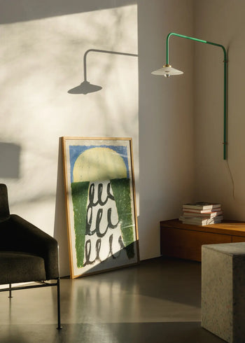 Sunlight fills a minimalist room with a modern black chair, The Poster Club Suzanne Lustig - Energize (30x40) art print leaning against the wall, books stacked on a wooden bench, and a green wall-mounted lamp.