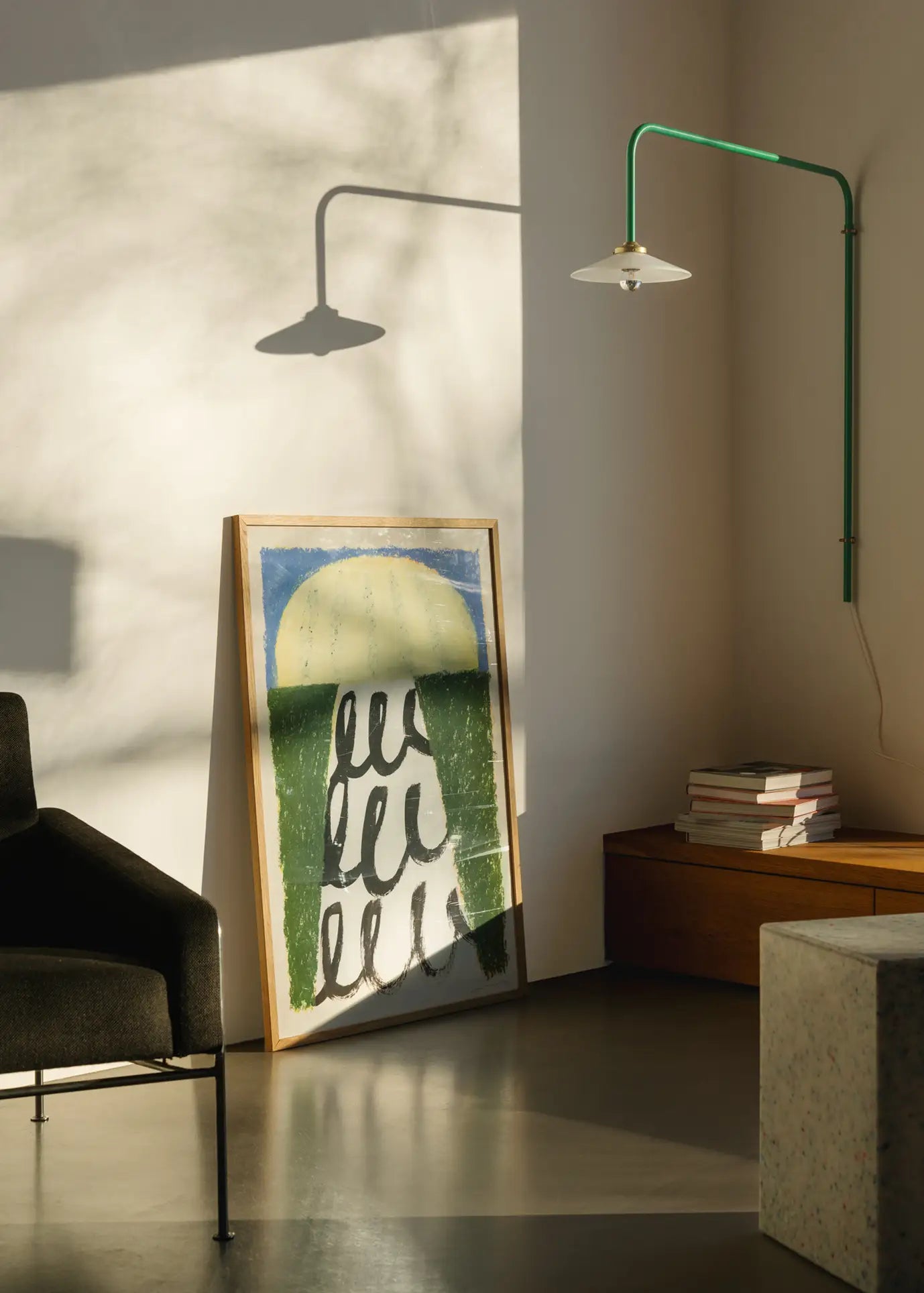 Sunlight fills a minimalist room where The Poster Clubs Suzanne Lustig - Energize (50x70) art print rests beside books, a black chair, and a green wall-mounted lamp.