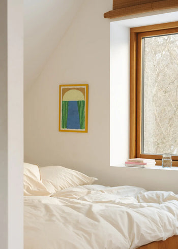 A cozy bedroom featuring white bedding, The Poster Club Suzanne Lustig - Laughing Jim (50x70) print in green, blue, and yellow tones on the wall, a wooden-framed window, several books, and a glass of water on the windowsill.