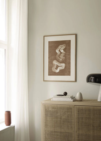 A minimalist room features a rattan cabinet, books, and decor beneath The Poster Club Tenna Elisabeth Studio - Lineshape 03 (50x70) art print. Natural light filters through large windows with sheer white curtains.