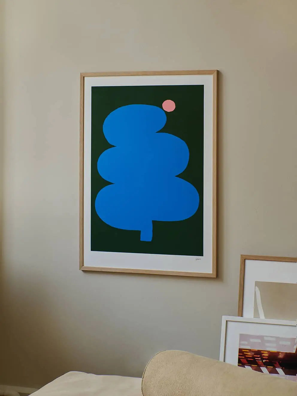 The Poster Club Wensi Zhai - Fat Tree Pink Moon (50x70), featuring an abstract blue tree and pink circle on a dark green background, is framed and hangs above other fine art prints leaning on a beige wall.