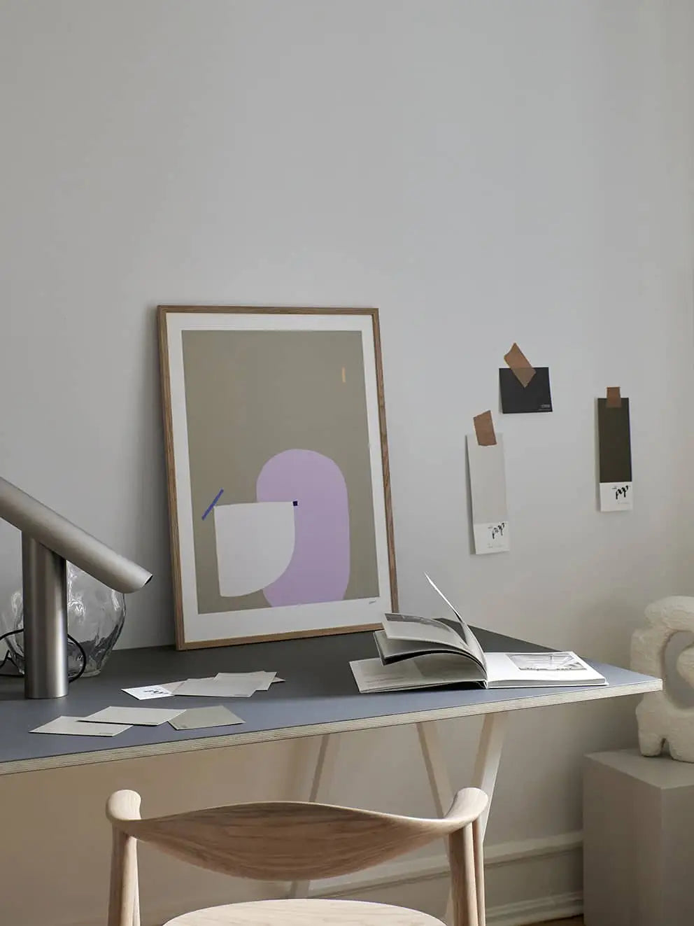 A modern, minimal desk setup features The Poster Club Wensi Zhai - I Like Shapes 08 (50x70) print against a white wall, paint swatches nearby, an open book on the desk, and a wooden chair in the foreground.