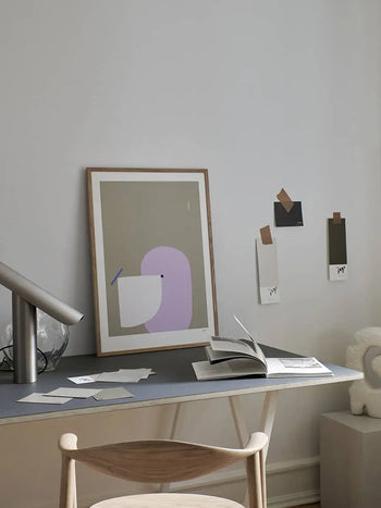 A modern, minimal desk setup features The Poster Club Wensi Zhai - I Like Shapes 08 (50x70) print against a white wall, paint swatches nearby, an open book on the desk, and a wooden chair in the foreground.