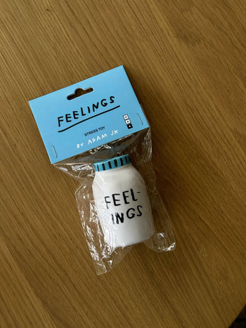 The Third Drawer Down Adam JK - Feelings Stress Toy, by Third Drawer Down, is a white pill bottle-shaped stress toy labeled FEELINGS, designed to help release emotions. Its packaged in clear plastic with a blue FEELINGS STRESS TOY BY ADAM JK tag.