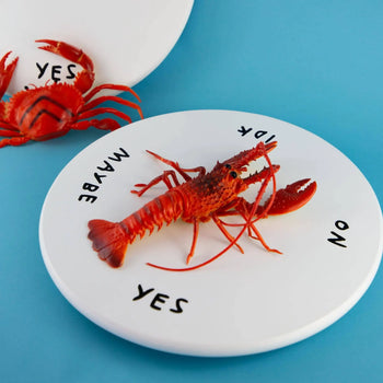 A red toy lobster rests on the Third Drawer Down Adam JK - Indecisive Lazy Susan plate, featuring YES, NO, MAYBE, and IDK around the edge. Another lobster and part of a plate sit in the background on a blue surface.