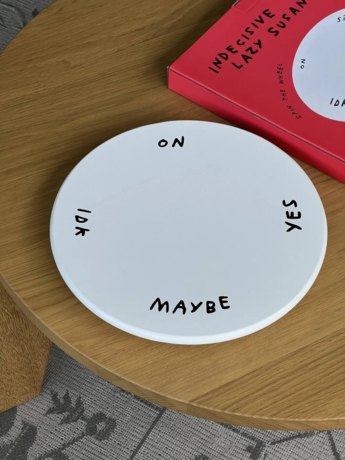 The Third Drawer Down Adam JK - Indecisive Lazy Susan is a white circular board marked ON, YES, MAYBE, and IDK. Sitting by its red box, this playful product from Third Drawer Down offers a fun way to make decisions when youre unsure.