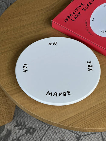 The Third Drawer Down Adam JK - Indecisive Lazy Susan is a white circular board marked ON, YES, MAYBE, and IDK. Sitting by its red box, this playful product from Third Drawer Down offers a fun way to make decisions when youre unsure.
