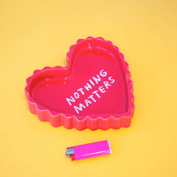 A heart-shaped pink ceramic tray by Third Drawer Down, reading NOTHING MATTERS, sits next to a pink lighter on a bright yellow background.