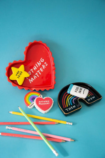 The Third Drawer Down Adam JK - Nothing Matters Ceramic Tray features playful shapes and phrases like “NOTHING MATTERS” and “YOU TRIED,” adding a whimsical touch to any space. Displayed on a blue surface with pencils and erasers nearby.