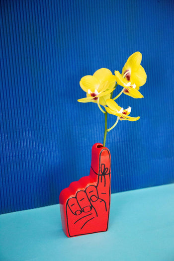 The Third Drawer Down Adam JK - Remember Bud Ceramic Vase, shaped like a red foam finger and filled with yellow orchids, stands out against a blue textured backdrop and light blue surface.