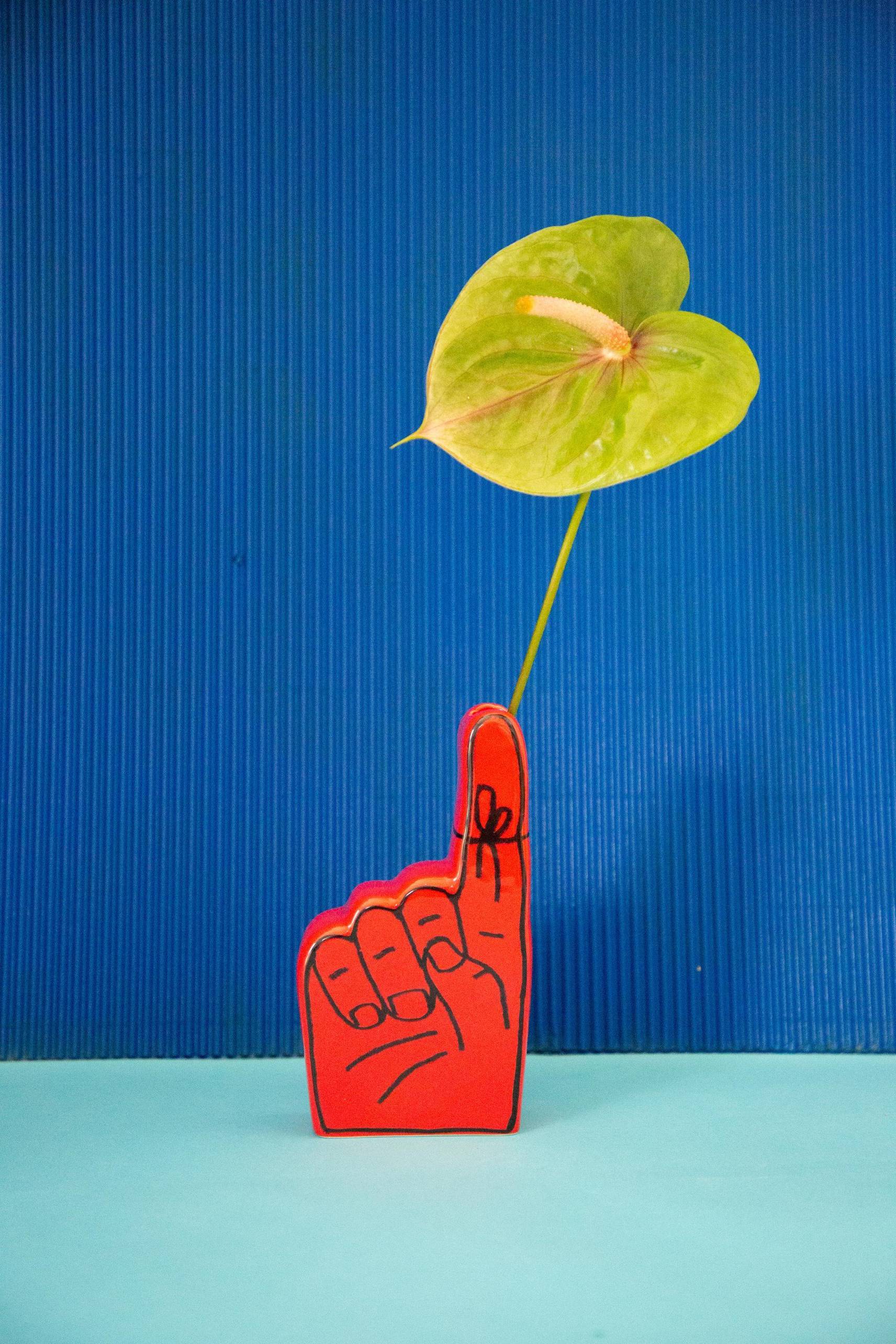 A green anthurium stands tall in the Third Drawer Down Adam JK - Remember Bud Ceramic Vase, with a red foam finger pointing up, set against a blue background and light blue surface.