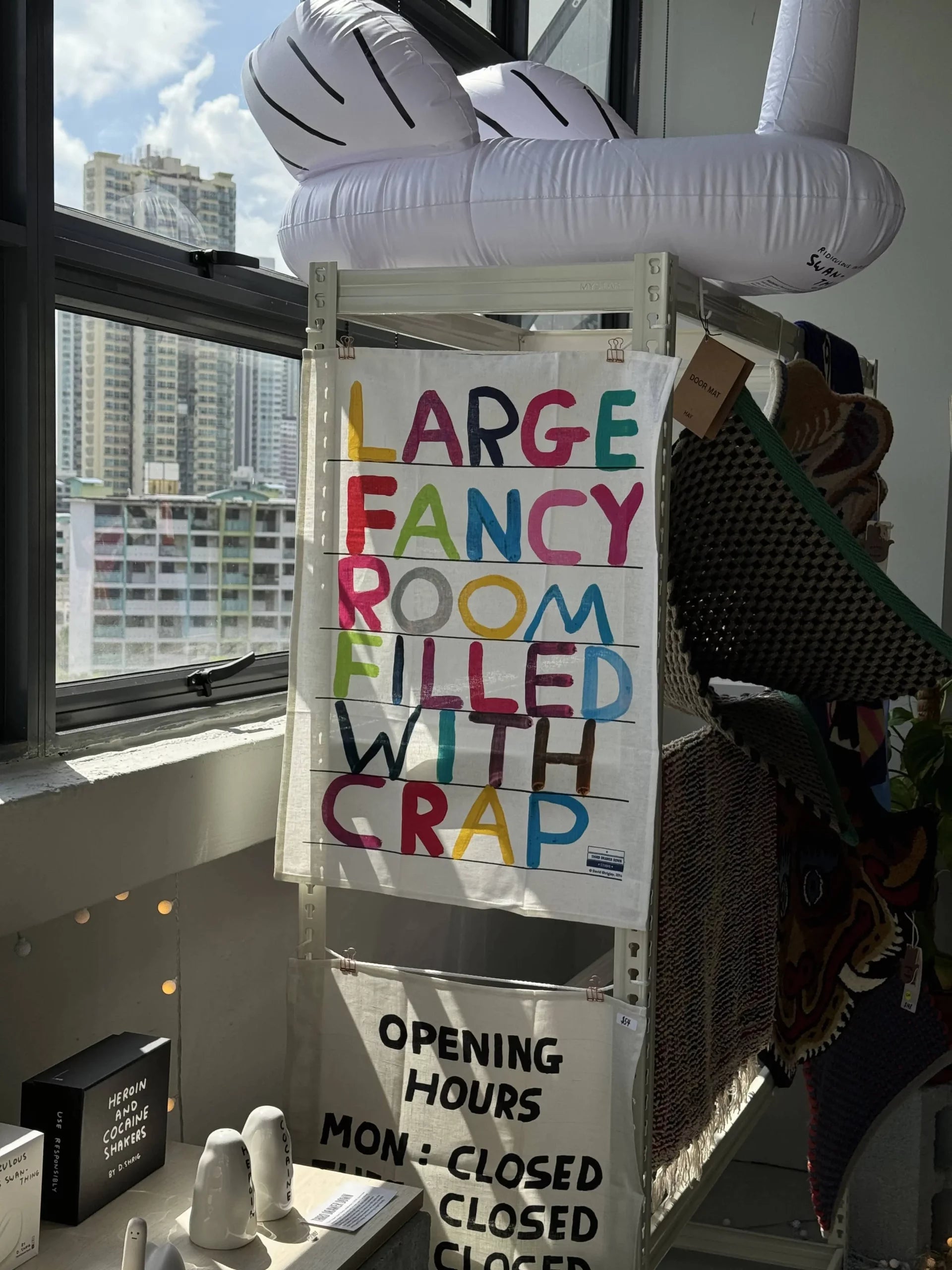 A colorful tea towel by Third Drawer Down displays LARGE FANCY ROOM FILLED WITH CRAP, seen near a window with city views—an ideal gift for any David Shrigley fan.