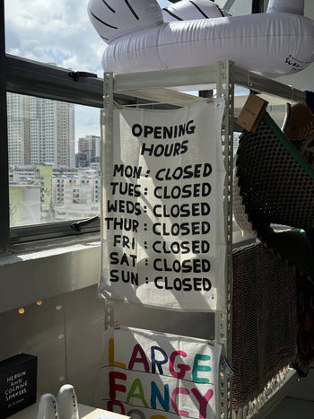 The Third Drawer Down David Shrigley - Opening Hours Tea Towel by Third Drawer Down hangs on a shelf in a sunlit store, each day marked CLOSED, with city buildings seen through the window behind.
