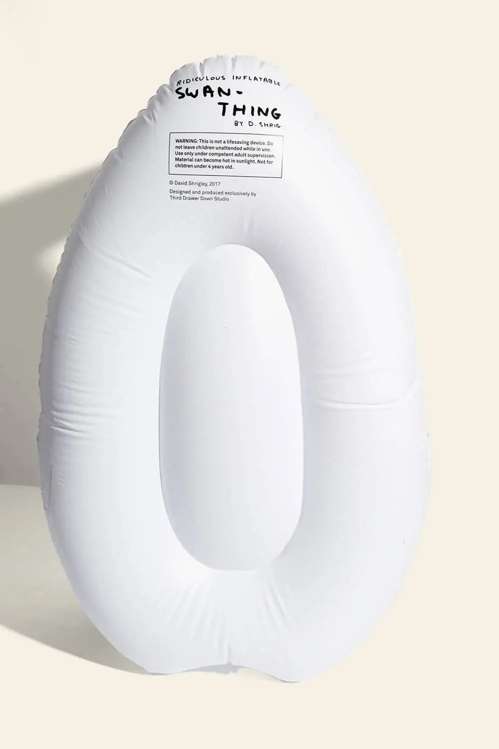 A white, oval inflatable pool float from Third Drawer Down stands upright against a beige background. Black text at the top reads “SWAN THING”—a playful reference to the Third Drawer Down David Shrigley Ridiculous Inflatable Swan-Thing.
