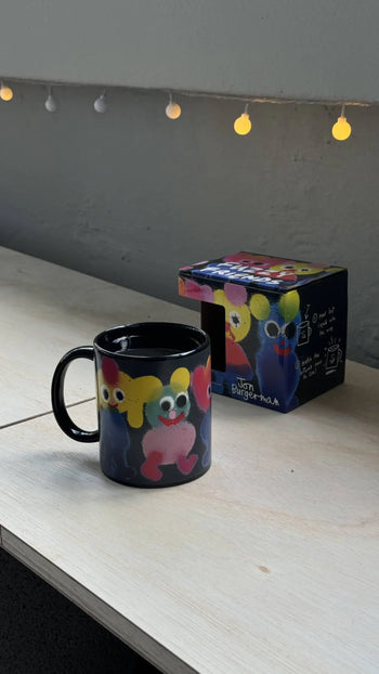 The Third Drawer Down Jon Burgerman Fuzzy Friend Mug, featuring colorful cartoon creatures, sits on a light wooden table with its matching box behind it and soft string lights glowing above in the background.