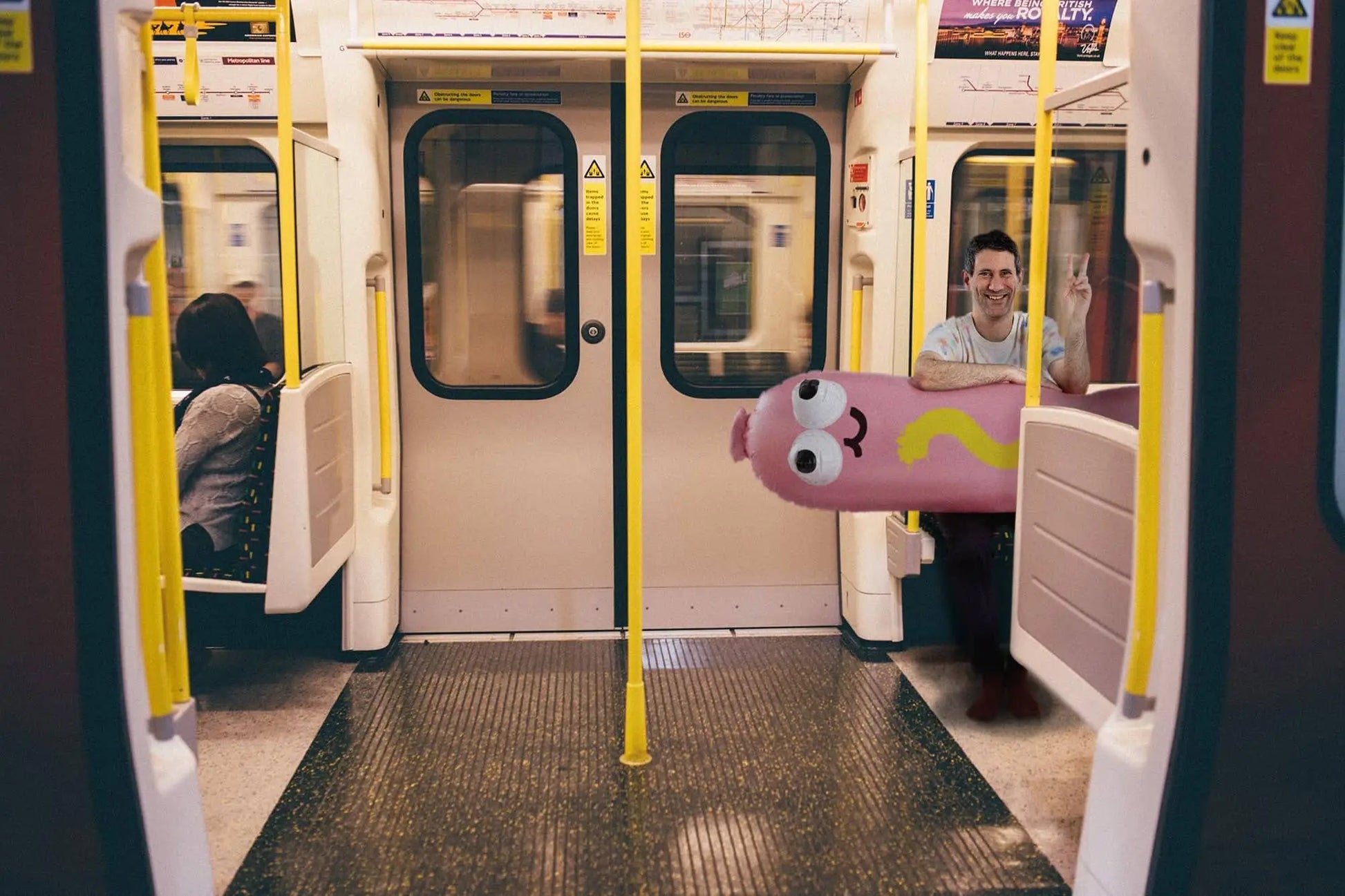 A man smiles on a subway, holding the Third Drawer Down Jon Burgerman - Hot Diggity Dog Pool Float, a large, colorful inflatable shaped like a cartoon worm with googly eyes. Another person sits farther away in the mostly empty car.