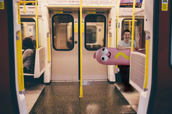 A man smiles on a subway, holding the Third Drawer Down Jon Burgerman - Hot Diggity Dog Pool Float, a large, colorful inflatable shaped like a cartoon worm with googly eyes. Another person sits farther away in the mostly empty car.