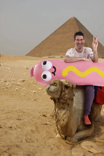 A smiling man sits on a camel in front of a pyramid, flashing a peace sign while holding the Third Drawer Down Jon Burgerman - Hot Diggity Dog Pool Float, a playful inflatable by Third Drawer Down perfect for summer pool parties.