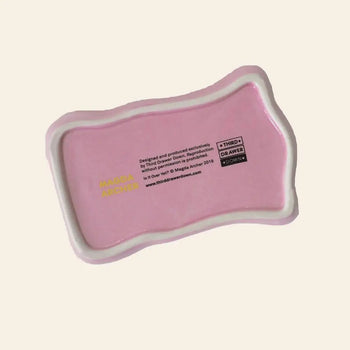 The Third Drawer Down Magda Archer - Is It Over Yet Trinket Tray is a pink, irregular ceramic tray with a white border and black/yellow text—ideal for small jewelry—by Third Drawer Down, shown on a light beige background.