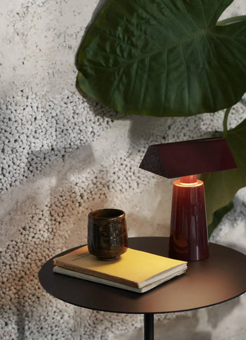 A round black side table holds a yellow notebook, a dark ceramic cup, and the &Tradition Caret MF1 Portable Table Lamp by &Tradition. Behind the table, a large green leaf rests against a textured light gray wall.