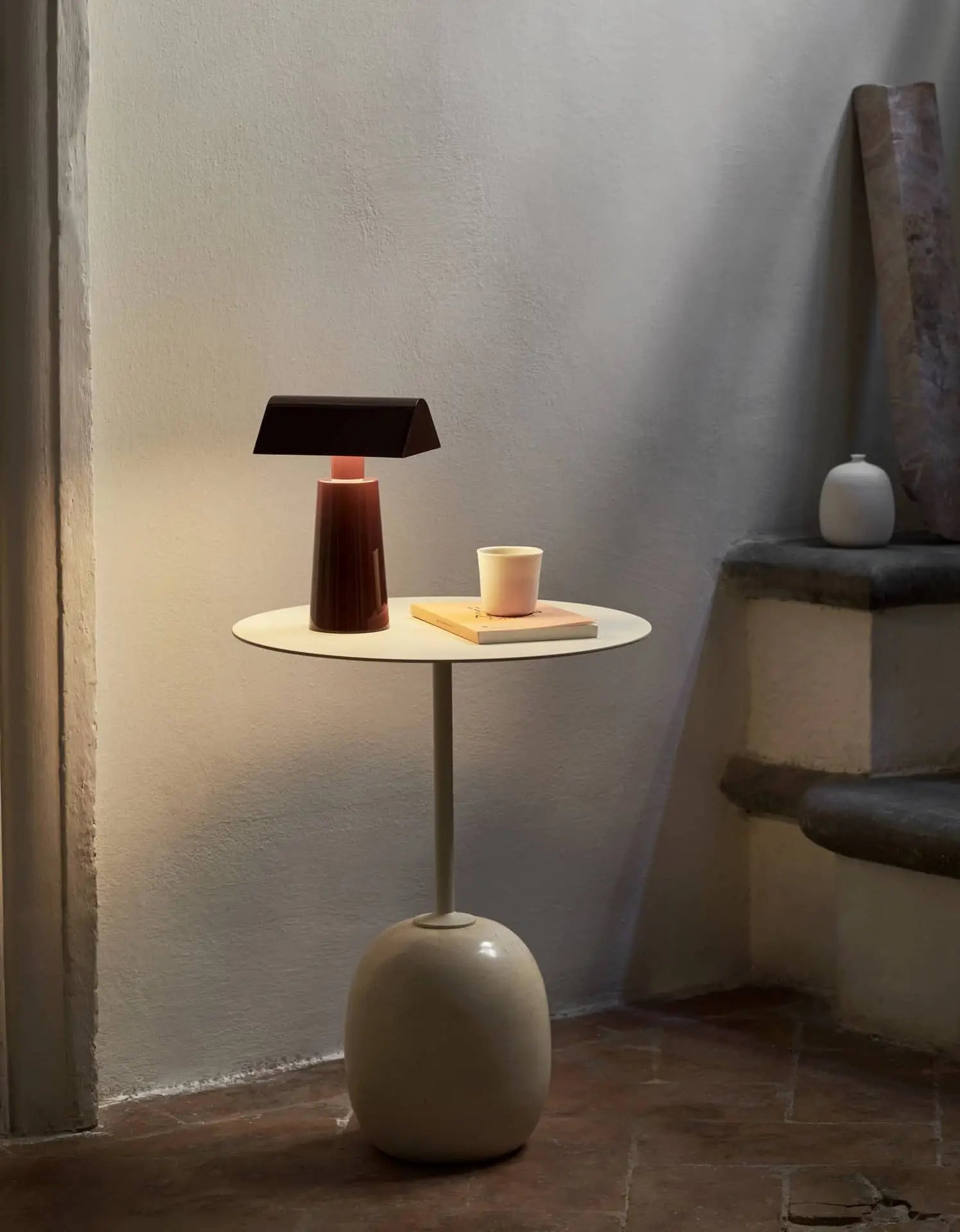 A modern round side table holds an &Tradition Caret MF1 Portable Table Lamp in sleek red, a book, and a cup. Set by stone steps against a light wall, it’s accented with a small white vase on the step for added charm.