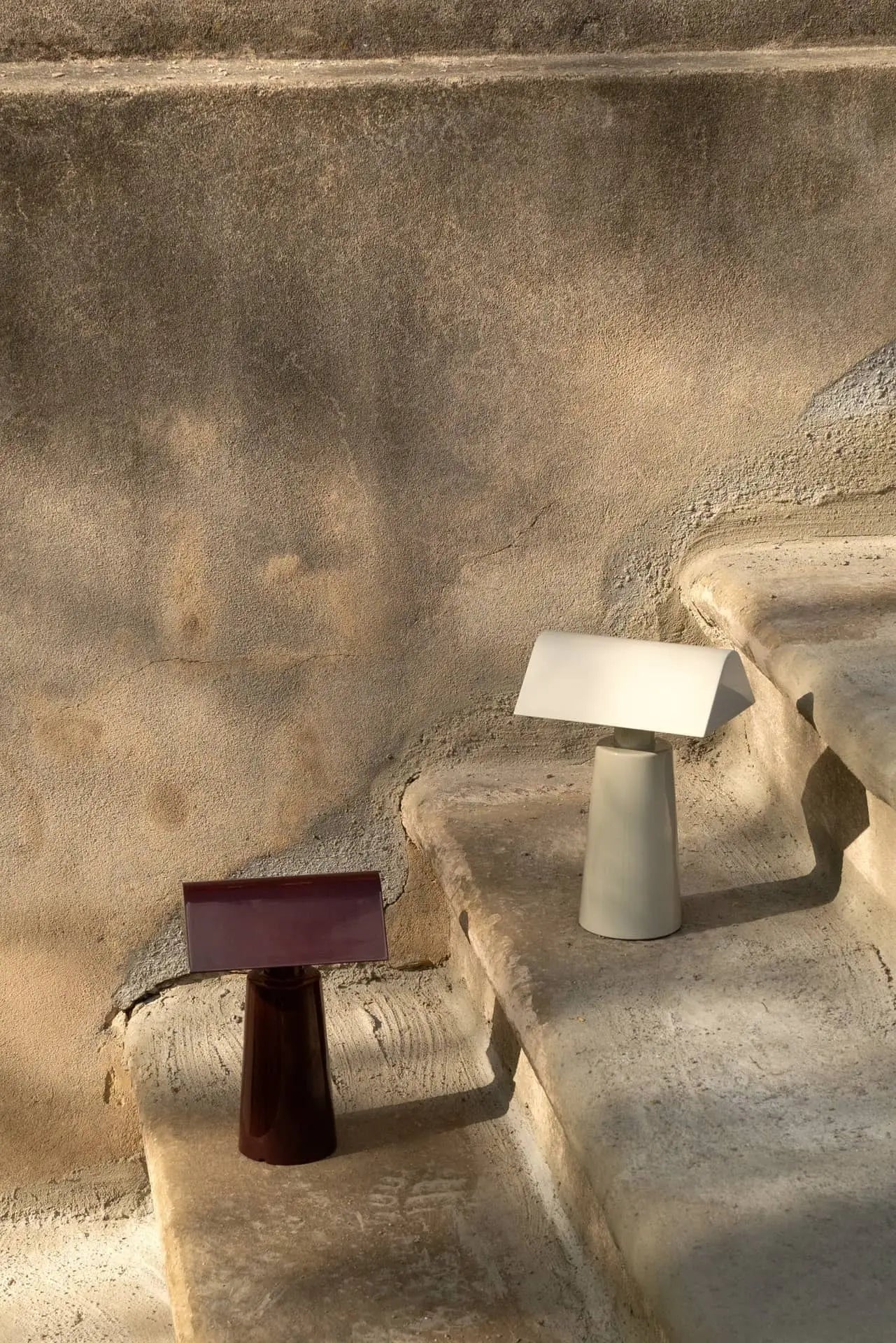 Two &Tradition Caret MF1 Portable Table Lamps, one white and one burgundy, by &Tradition, rest on sunlit stone steps against a weathered beige wall, with light and shadow enhancing their modern minimalist design.