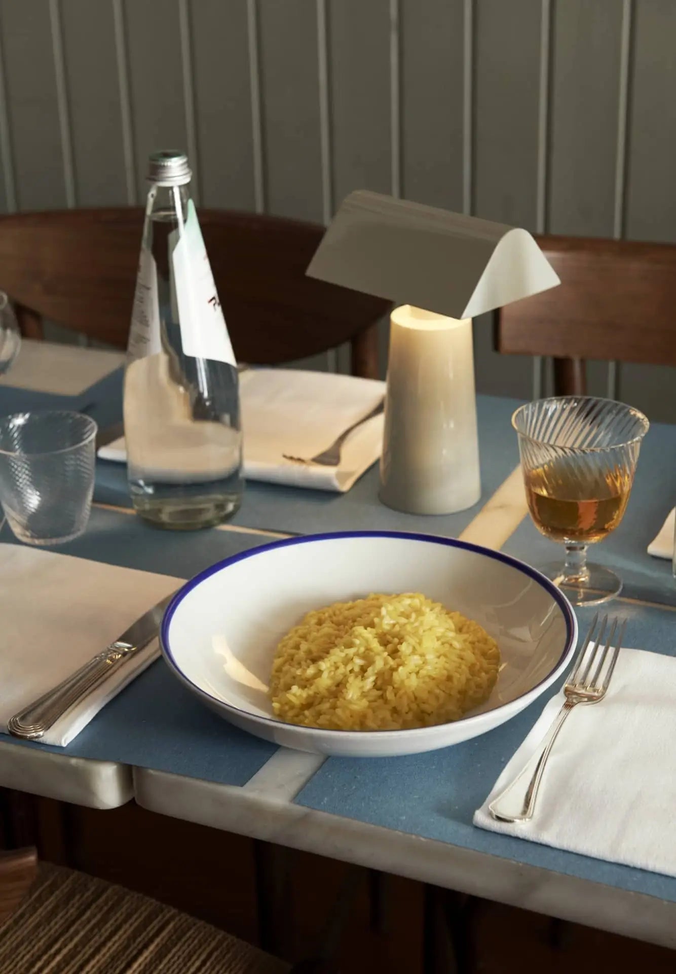 A bowl of yellow risotto sits on a set dining table with a water bottle, amber drink, utensils, napkins, glasses, and an &Tradition Caret MF1 Portable Table Lamp by &Tradition featuring modern design.