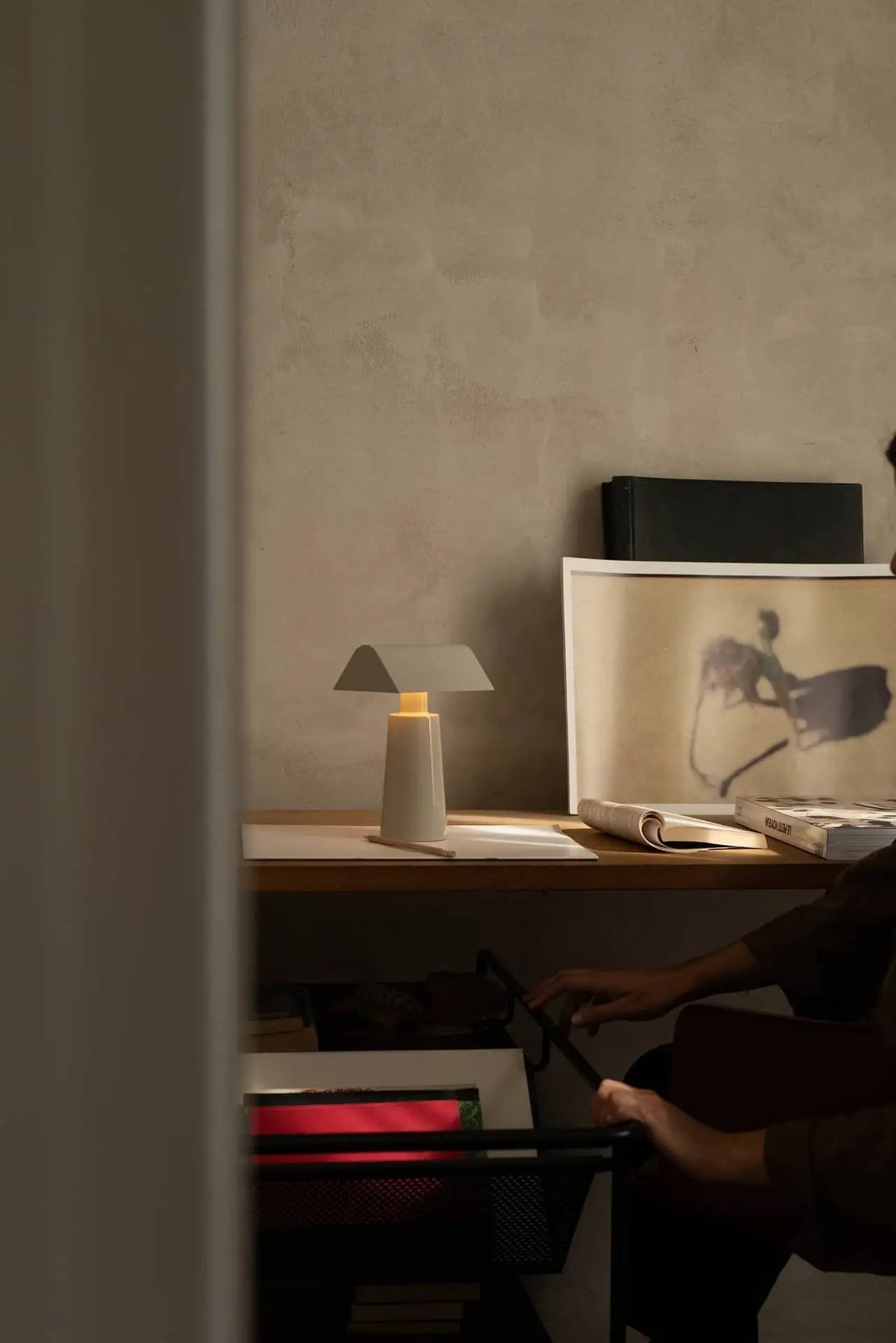 A dimly lit desk features the &Tradition Caret MF1 Portable Table Lamp by &Tradition, books, and horse-and-rider shadow art. A partially visible person reaches toward a dark cart beneath the desk.