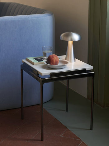 A small marble-topped side table holds a plate with a peach, a glass of water, a book, and the &Tradition Como SC53 Portable Table Lamp. The table sits beside a light blue sofa and against a green wall.