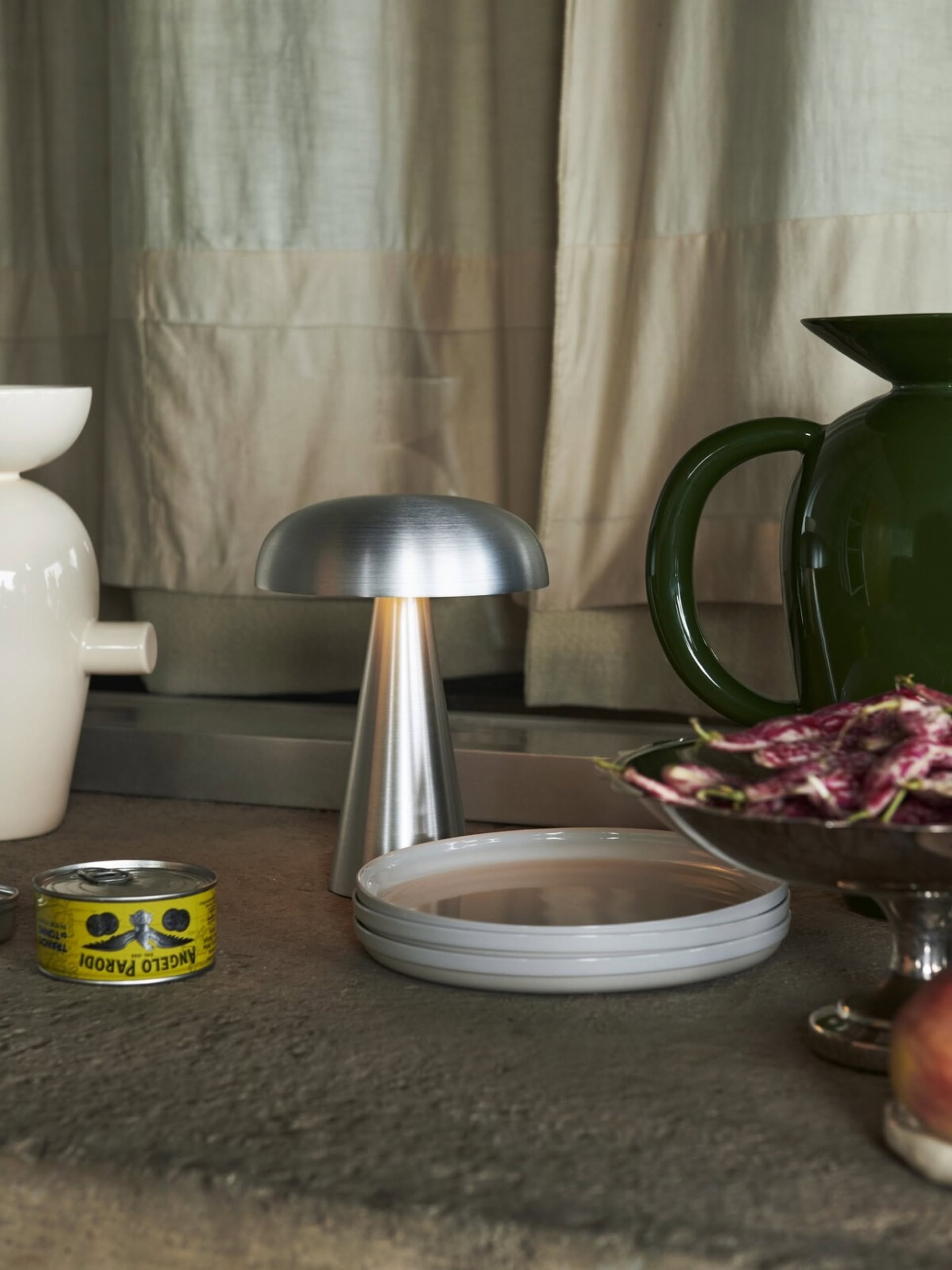 An &Tradition Como SC53 Portable Table Lamp sits on a stone surface near stacked white plates, a green pitcher, a can of anchovies, a white vase, and a dish of radicchio, with cream curtains in the background.