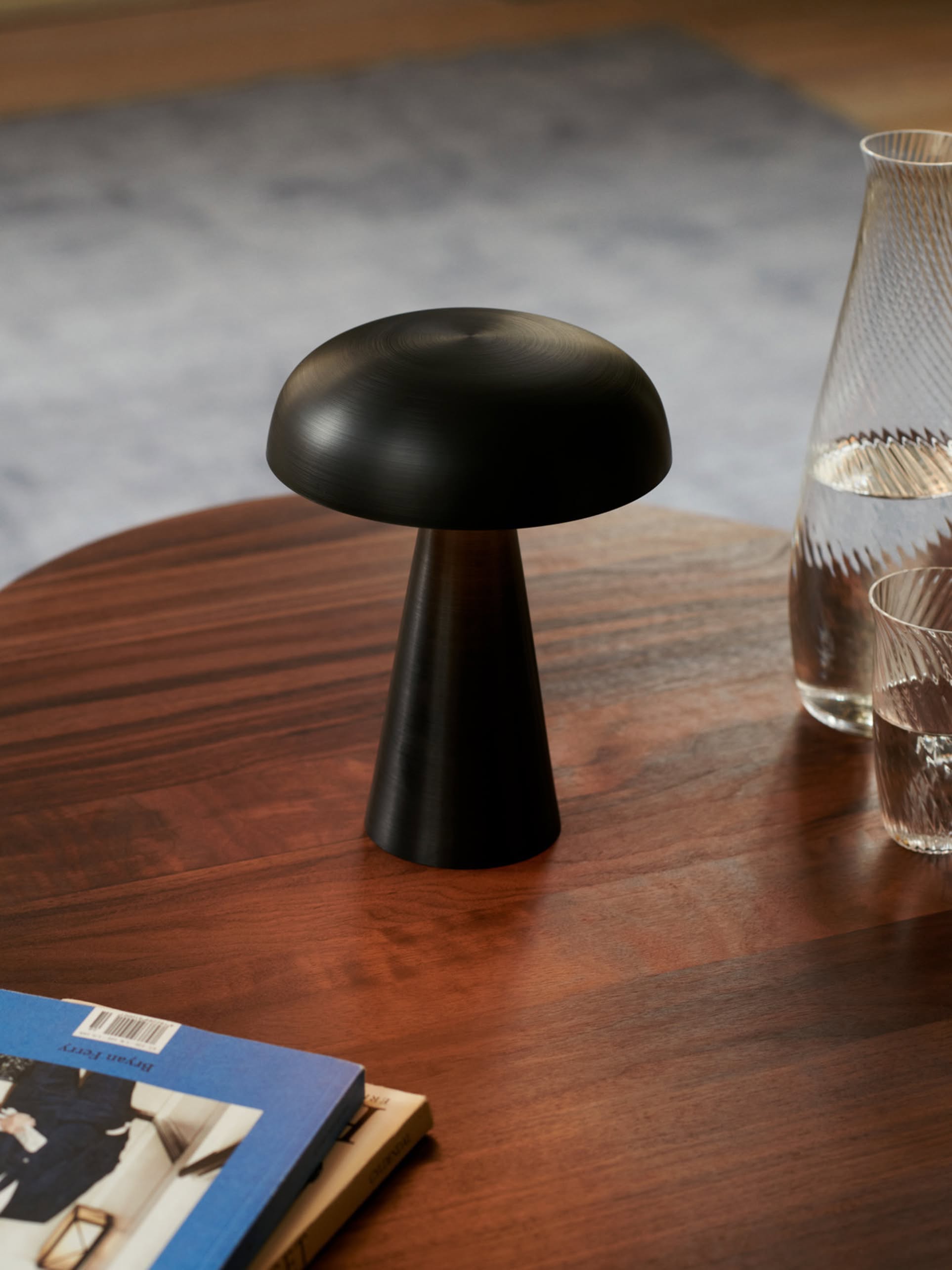 The &Tradition Como SC53 Portable Table Lamp by &Tradition, with its modern black mushroom shape, sits on a wooden table beside a glass pitcher, drinking glass, and a blue book; a blurred carpet is seen in the background.