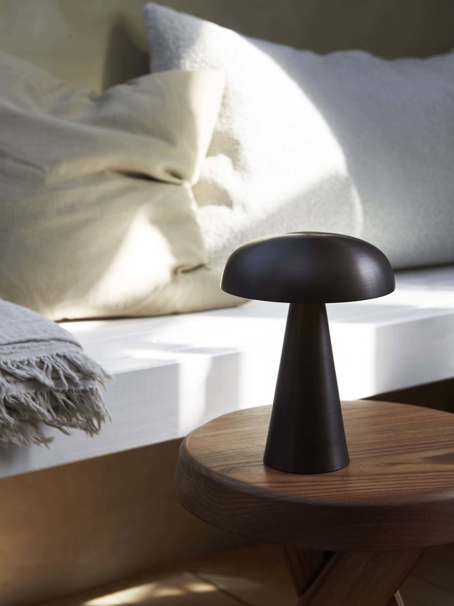 The &Tradition Como SC53 Portable Table Lamp, a small modern black mushroom-shaped lamp by &Tradition, sits on a wooden table next to a bench with light textured cushions and a fringed throw blanket in soft natural daylight.