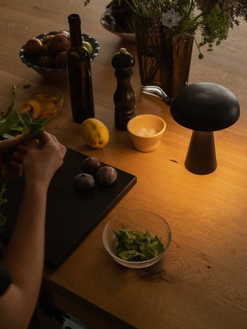 A person prepares food at a wooden table adorned with figs, lemon, fresh greens, and more, all warmly lit by the &Tradition Como SC53 Portable Table Lamp—an adjustable light that complements the pepper grinder, bottle, salt bowl, and fruit.