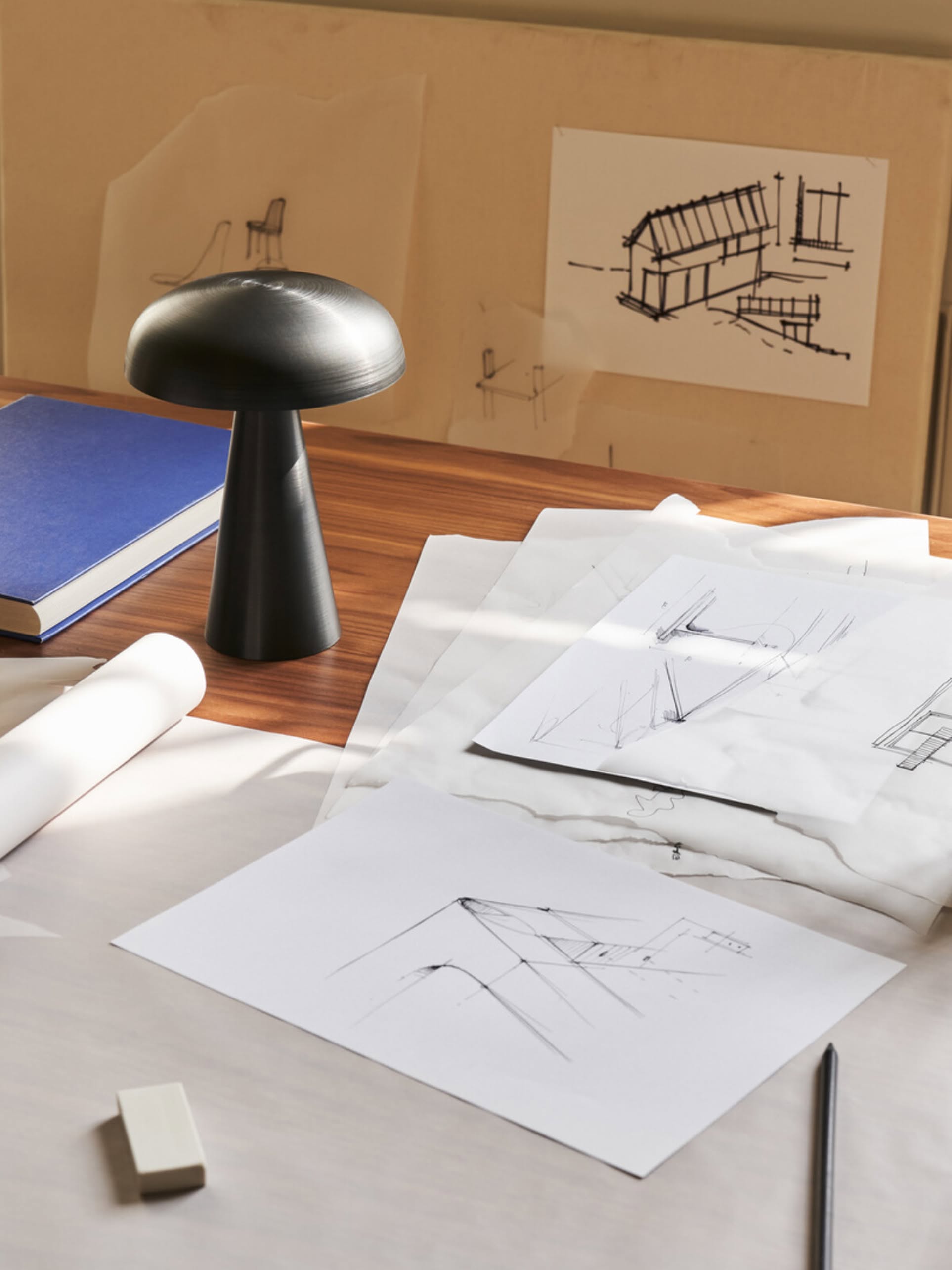 A wooden desk with architectural sketches, tracing paper, a rechargeable black &Tradition Como SC53 Portable Table Lamp with adjustable lighting, a pencil, an eraser, and a blue book. Drawings of buildings are pinned behind the desk.
