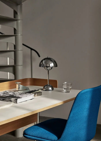 A modern workspace features a white desk, blue upholstered chair, USB cable, stacked books, glasses, a glass tumbler, and the &Tradition Flowerpot VP9 Portable Table Lamp by &Tradition. A spiral staircase is visible in the background.